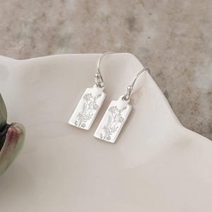 Puede incluir: Un par de pendientes colgantes rectangulares plateados con un diseño floral. Los pendientes se exhiben en un plato blanco de forma única, junto a una suculenta de cerámica verde y granate. El fondo es gris neutro.