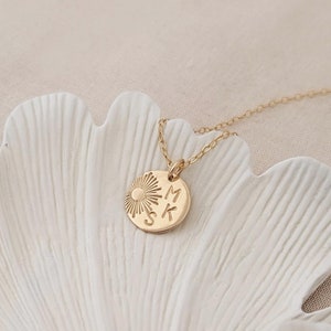 Puede incluir: Un collar de oro con un pequeño colgante redondo que presenta un sol y las letras "MSK". El collar se muestra sobre una hoja de ginkgo de cerámica blanca.