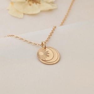 May include: A delicate gold-colored necklace featuring a pendant with sun and moon designs. The pendant hangs from a fine chain. The background includes dried flowers, creating a soft, natural aesthetic. The necklace is a piece of jewelry.