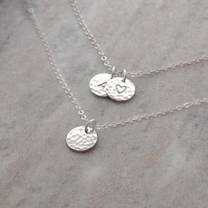 May include: Two silver necklaces with hammered-texture round pendants. One necklace has a single pendant, while the other has two pendants, one with the letters "A" and "V" and a heart symbol. The necklaces are displayed on a gray surface.
