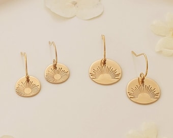 May include: Four pairs of gold-coloured earrings with a sunburst design are displayed on a white hexagonal tray with a gold rim. White flowers are scattered around the earrings, creating a delicate and elegant presentation.
