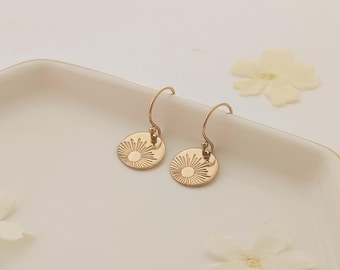 May include: Gold-coloured earrings with a sunburst design, displayed on a white tray. The earrings are small, circular, and have hook closures. The tray is white with a gold trim, decorated with white flowers.