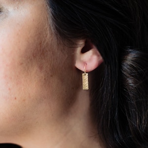 May include: A close-up of a woman's ear with a gold bar earring. The earring is textured and has a hammered finish.