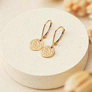 May include: A pair of gold-colored earrings with a hammered disc design, displayed on a white circular pedestal. The earrings have a simple, elegant design. The background features dried flowers.