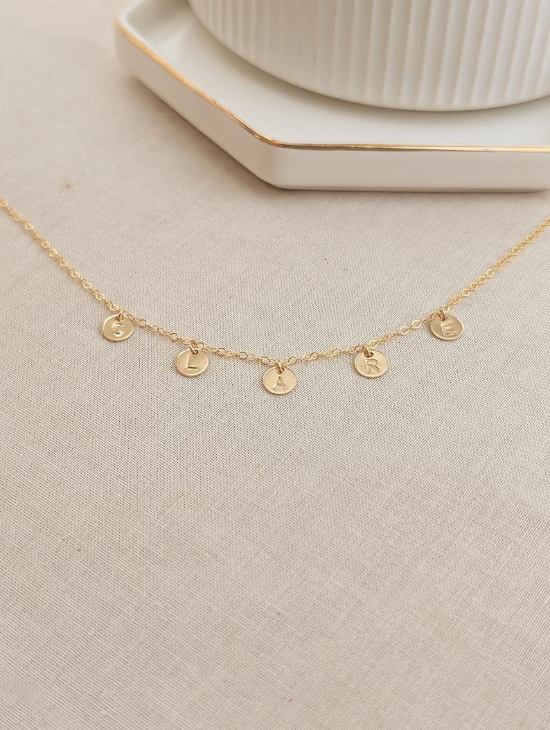 May include: A gold chain necklace with five small gold discs, each with a letter engraved on it. The letters spell out "GRACE".