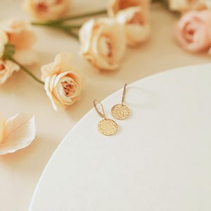 May include: A pair of gold-colored, hammered-texture disc earrings with a simple hook design. The earrings are displayed on a white surface, with soft-focus peach-colored roses in the background. The earrings are a delicate jewelry item.