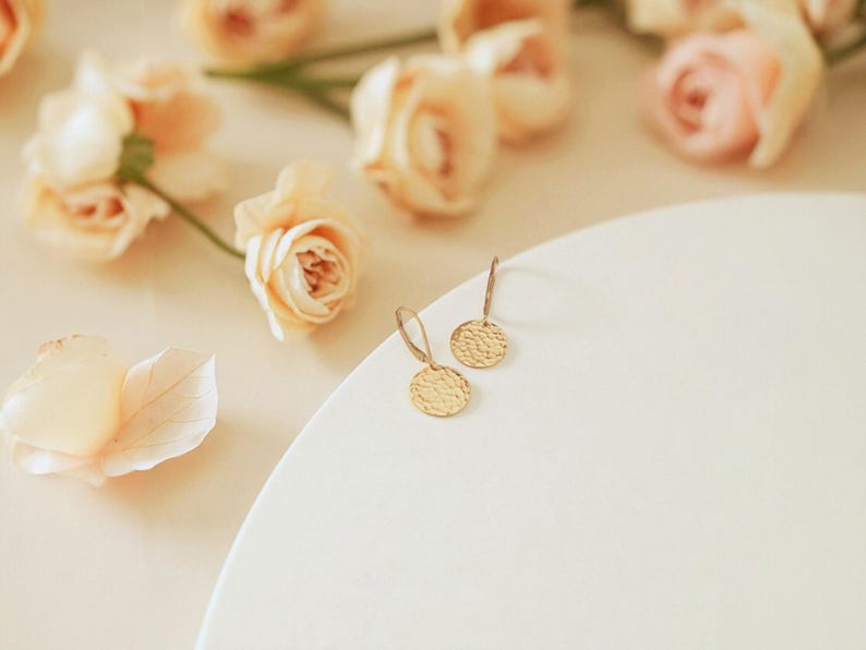 May include: A pair of gold-colored, hammered-texture disc earrings with a simple hook design. The earrings are displayed on a white surface, with soft-focus peach-colored roses in the background. The earrings are a delicate jewelry item.
