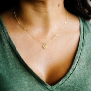 May include: A gold-colored necklace with a small, round pendant featuring a sun and moon design. The necklace is worn against a green v-neck shirt. The chain is delicate and the pendant is centered.