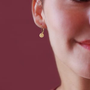 May include: A gold-colored earring with a hammered disc pendant is shown. The earring is a small hoop style, and the disc has a textured surface. The background is a solid maroon color.
