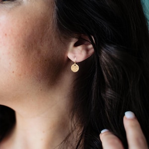 May include: A close-up of a gold-colored, textured disc earring hanging from a small hook. The earring is round with a hammered texture. The image shows the earring on a person's ear, with dark hair and skin visible.