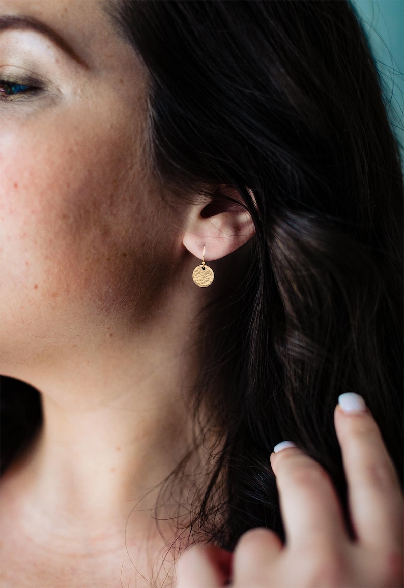 May include: A close-up of a gold-colored, textured disc earring hanging from a small hook. The earring is round with a hammered texture. The image shows the earring on a person's ear, with dark hair and skin visible.
