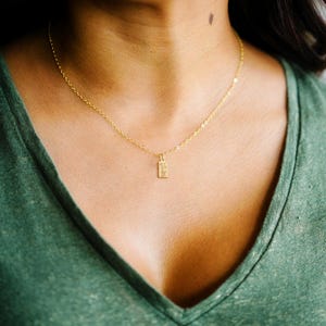 May include: A delicate gold necklace with a small rectangular pendant. The pendant has a textured surface and hangs from a fine chain. The necklace is worn against a green shirt.