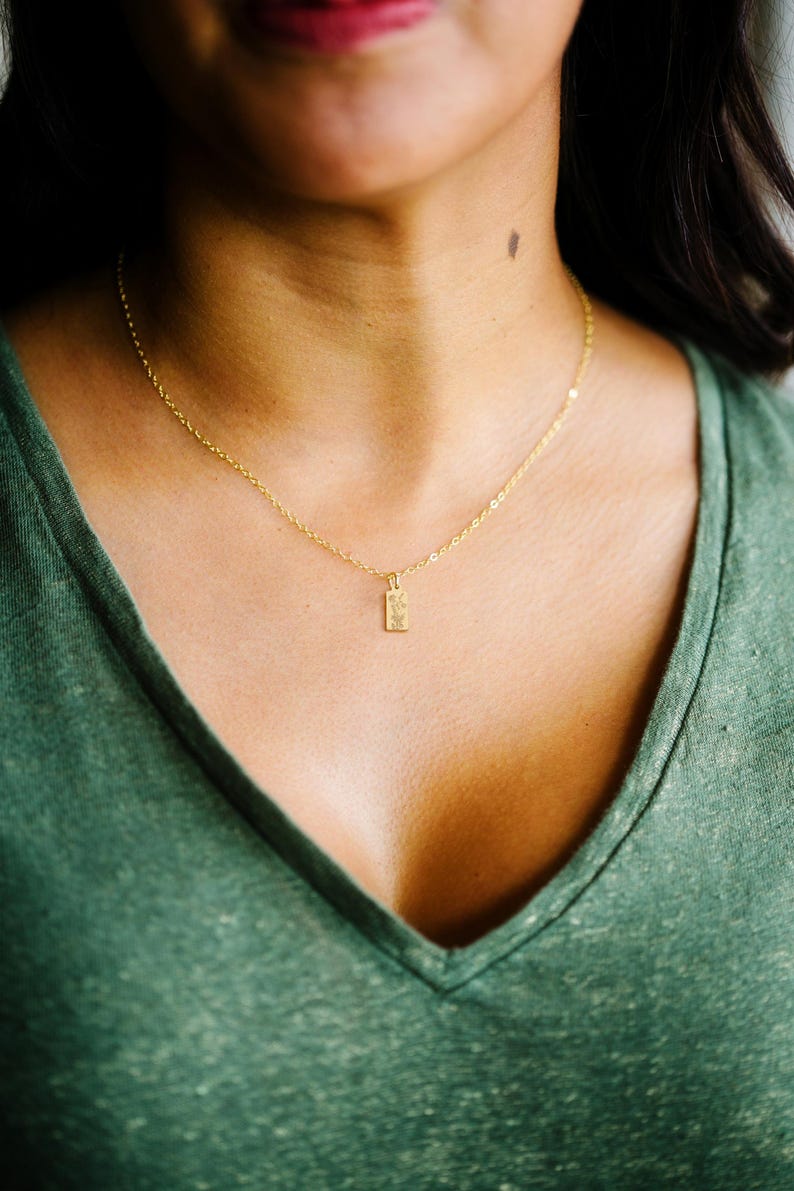 May include: A delicate gold necklace with a small rectangular pendant. The pendant has a textured surface and hangs from a fine chain. The necklace is worn against a green shirt.
