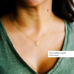 May include: A delicate gold necklace with a rectangular pendant. The pendant has a textured design. The model is wearing a green v-neck shirt. Text on the image says "Also available in gold in the shop!"