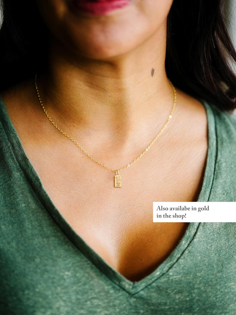 May include: A delicate gold necklace with a rectangular pendant. The pendant has a textured design. The model is wearing a green v-neck shirt. Text on the image says "Also available in gold in the shop!"