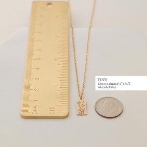 May include: A gold-colored necklace with a rectangular pendant featuring a floral design. The pendant measures 12mm x 6mm (&frac12;" x &frac14;"). The necklace is displayed with a ruler and a coin for size comparison. The text "TINY!" and "14k Gold Filled" are also visible.