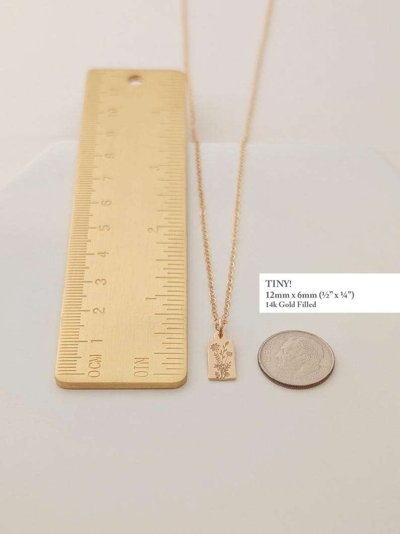 May include: A gold-colored necklace with a rectangular pendant featuring a floral design. The pendant measures 12mm x 6mm (&frac12;" x &frac14;"). The necklace is displayed with a ruler and a coin for size comparison. The text "TINY!" and "14k Gold Filled" are also visible.