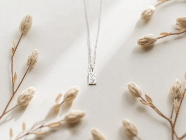 May include: A silver necklace with a rectangular pendant featuring a floral design. The pendant hangs from a delicate chain. Dried, beige-colored flowers are arranged around the necklace on a white surface, creating a minimalist aesthetic.