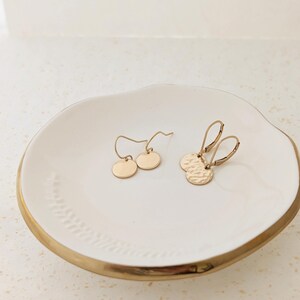 May include: Two pairs of gold-colored earrings displayed on a white ceramic dish with a gold rim. One pair features smooth, round discs, while the other has hammered texture. The earrings are delicate and minimalist.