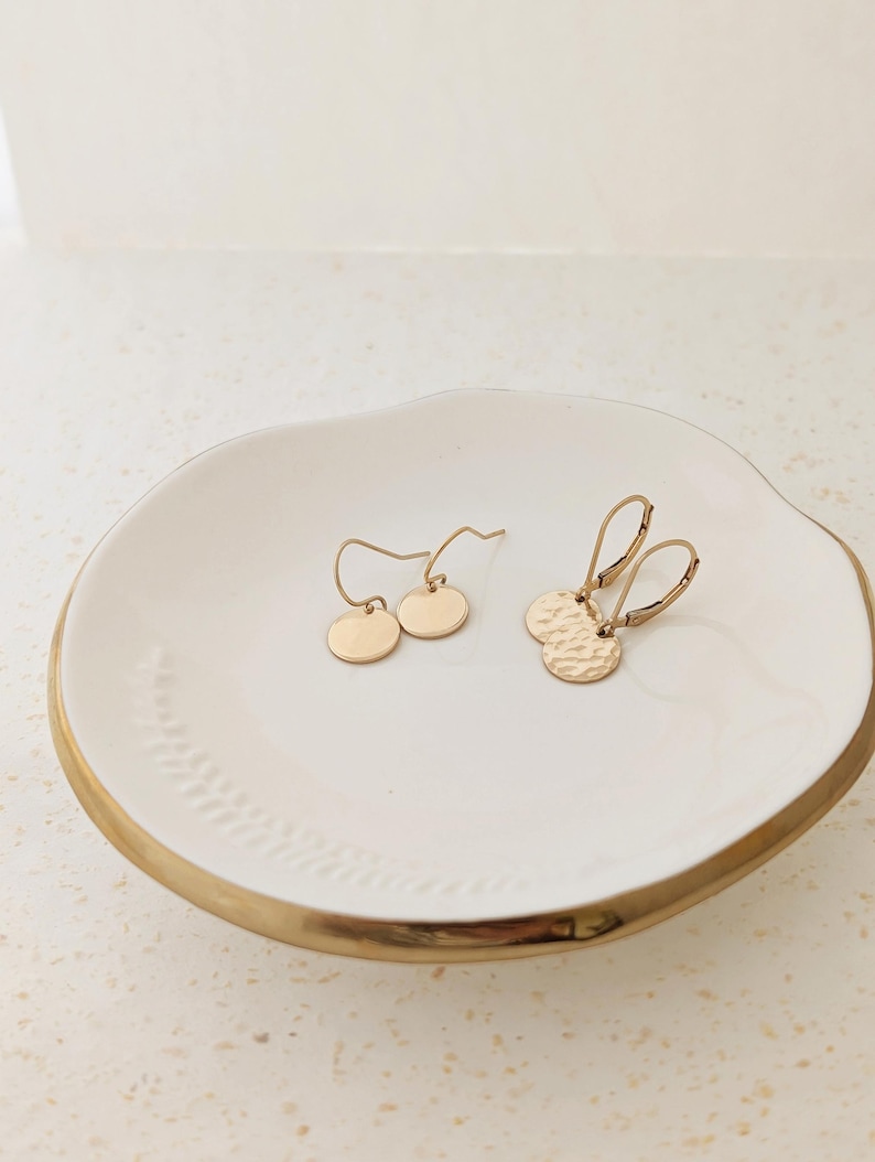 May include: Two pairs of gold-colored earrings displayed on a white ceramic dish with a gold rim. One pair features smooth, round discs, while the other has hammered texture. The earrings are delicate and minimalist.