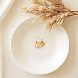 May include: A pair of gold-colored, textured disc earrings with curved ear wires, displayed on a white plate. The earrings are the focal point, with dried floral accents and a white cloth in the background. The earrings are approximately 0.75 inches in diameter.