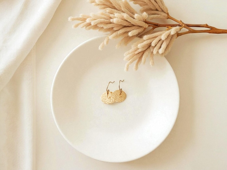 May include: A pair of gold-colored, textured disc earrings with curved ear wires, displayed on a white plate. The earrings are the focal point, with dried floral accents and a white cloth in the background. The earrings are approximately 0.75 inches in diameter.