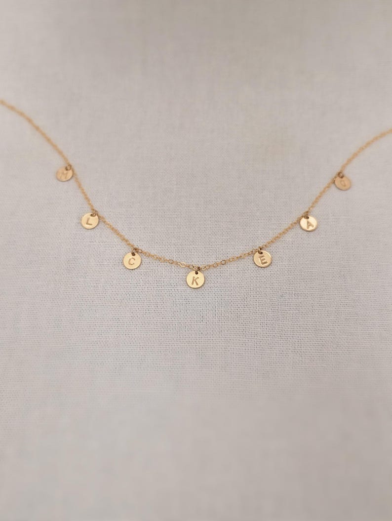 May include: A gold chain necklace with five small gold discs hanging from it. Each disc has a letter stamped on it, spelling out the word 'LAKE'. The necklace is on a white fabric background.