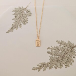 May include: A gold-colored necklace with a rectangular pendant featuring a floral design. The chain is delicate, and the pendant hangs in the center. The background includes dried fern leaves, creating a natural aesthetic.