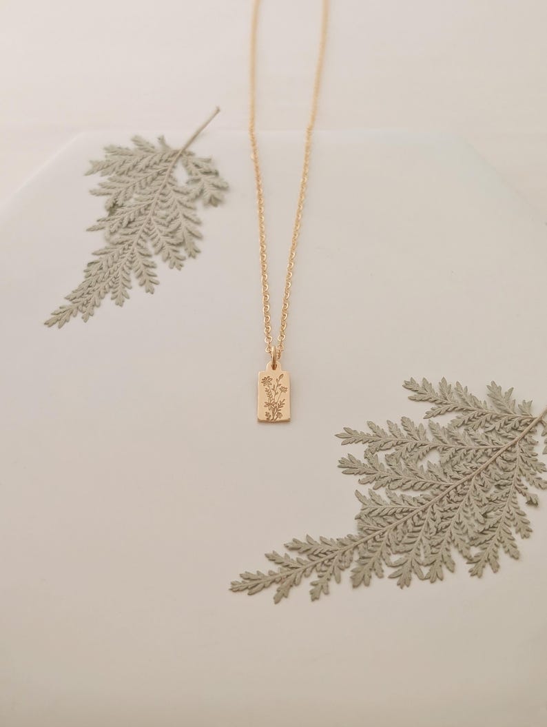 May include: A gold-colored necklace with a rectangular pendant featuring a floral design. The chain is delicate, and the pendant hangs in the center. The background includes dried fern leaves, creating a natural aesthetic.