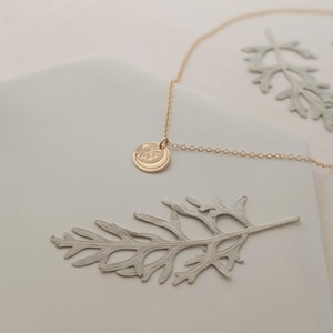 May include: A gold-colored necklace with a pendant featuring a sun and crescent moon design. The necklace is displayed on a white surface with two gray leaf-shaped decorations. The chain is delicate and the pendant is circular.