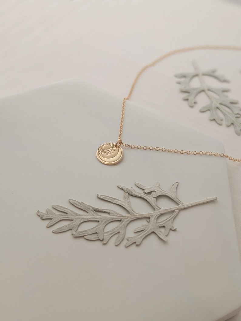 May include: A gold-colored necklace with a pendant featuring a sun and crescent moon design. The necklace is displayed on a white surface with two gray leaf-shaped decorations. The chain is delicate and the pendant is circular.