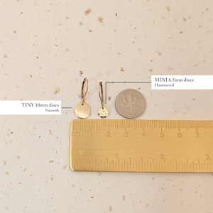 May include: A close-up of two gold-colored disc earrings, one smooth and 10mm, the other hammered and 6.5mm. A ruler and a dime are placed next to the earrings for size comparison. The text on the image reads "TINY 10mm discs Smooth" and "MINI 6.5mm discs Hammered".