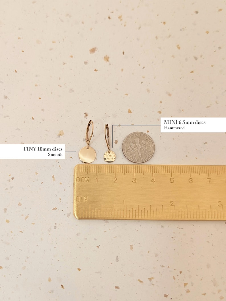May include: A close-up of two gold-colored disc earrings, one smooth and 10mm, the other hammered and 6.5mm. A ruler and a dime are placed next to the earrings for size comparison. The text on the image reads "TINY 10mm discs Smooth" and "MINI 6.5mm discs Hammered".