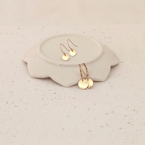 May include: Gold-colored disc earrings displayed on a white, petal-shaped dish. The earrings include a pair of small, simple drop earrings and a pair of hoop earrings with disc charms. The jewelry is set against a neutral background.