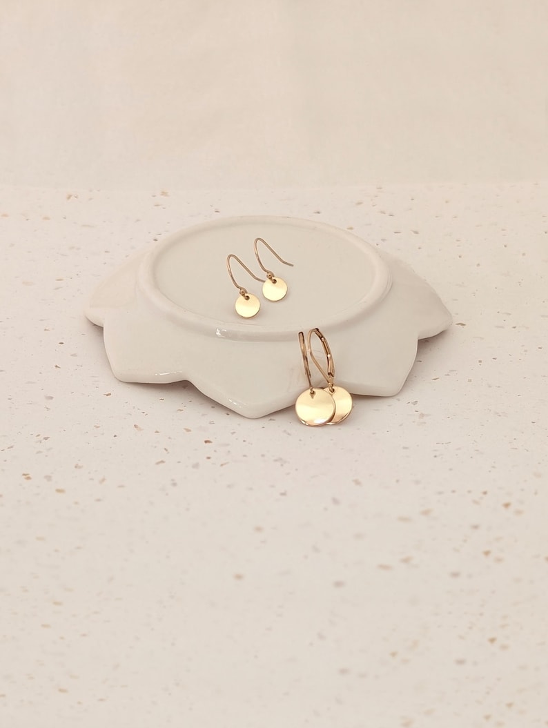 May include: Gold-colored disc earrings displayed on a white, petal-shaped dish. The earrings include a pair of small, simple drop earrings and a pair of hoop earrings with disc charms. The jewelry is set against a neutral background.