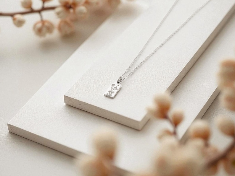 May include: A silver necklace with a rectangular pendant featuring a floral design. The pendant hangs from a delicate chain. The necklace is displayed on a white, layered surface, with soft, blurred floral accents in the foreground.