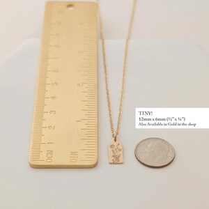 May include: A gold-colored necklace with a rectangular pendant featuring a floral design. The pendant measures 12mm x 6mm (&frac12;" x &frac14;"). The image includes a ruler and a coin for size comparison. The text "TINY!" is visible.