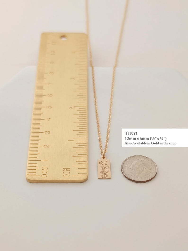 May include: A gold-colored necklace with a rectangular pendant featuring a floral design. The pendant measures 12mm x 6mm (&frac12;" x &frac14;"). The image includes a ruler and a coin for size comparison. The text "TINY!" is visible.