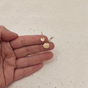 May include: Gold-colored disc earrings. One pair features a small, round stud, while the other has a larger disc suspended from a hook. The earrings are displayed against a neutral background, highlighting their simple, elegant design.