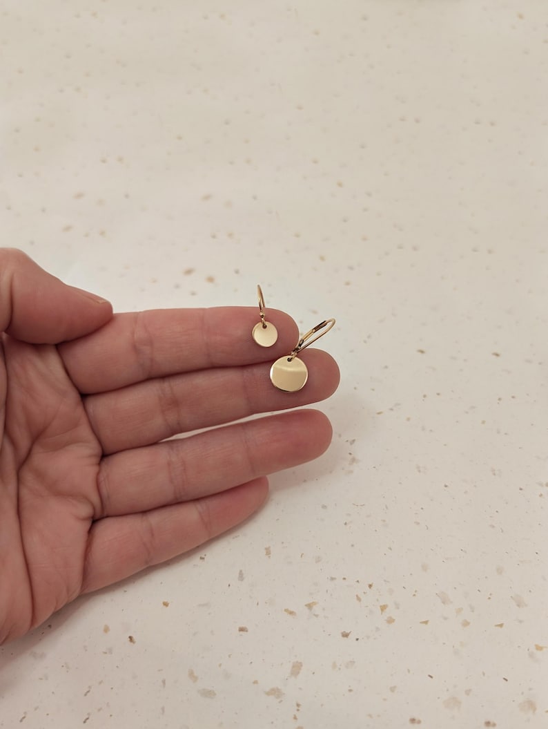 May include: Gold-colored disc earrings. One pair features a small, round stud, while the other has a larger disc suspended from a hook. The earrings are displayed against a neutral background, highlighting their simple, elegant design.
