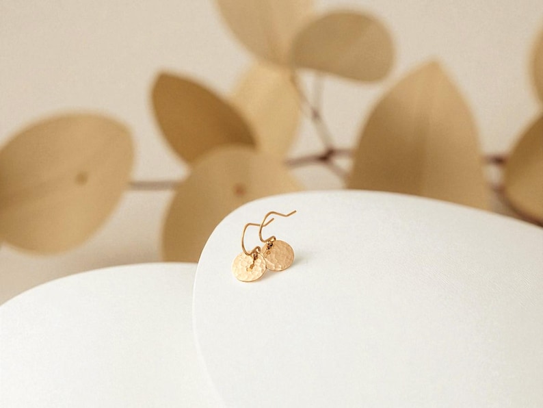 May include: A pair of gold-colored hammered disc earrings. The earrings are on a white surface, with a blurred background of beige-colored leaves. The earrings have a simple, elegant design.