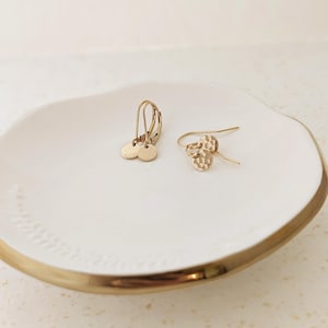 May include: Two pairs of gold-colored earrings on a white ceramic dish with a gold rim. One pair features small, round disc pendants, while the other has a textured, hammered design. The earrings are displayed on a white surface.