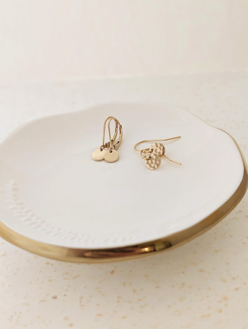 May include: Two pairs of gold-colored earrings on a white ceramic dish with a gold rim. One pair features small, round disc pendants, while the other has a textured, hammered design. The earrings are displayed on a white surface.