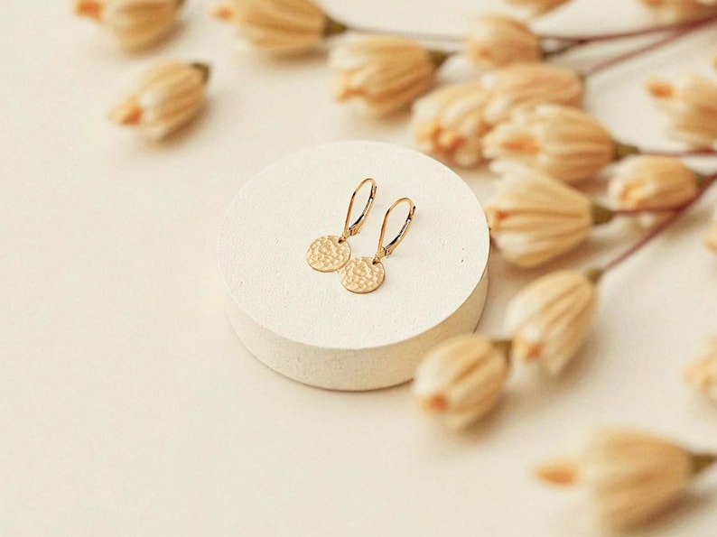 May include: A pair of gold-colored earrings with a hammered disc design, displayed on a white circular pedestal. The earrings have a simple, elegant design. The background features dried flowers.