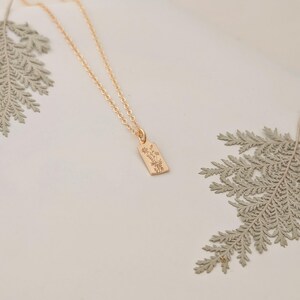 May include: A gold-colored necklace with a rectangular pendant featuring an engraved floral design. The necklace chain is delicate, and the pendant hangs from it. The background is a soft white, with fern leaves in the corners.