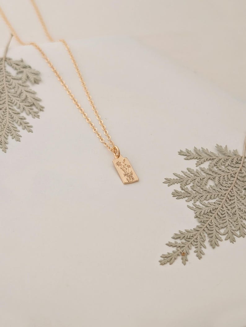 May include: A gold-colored necklace with a rectangular pendant featuring an engraved floral design. The necklace chain is delicate, and the pendant hangs from it. The background is a soft white, with fern leaves in the corners.