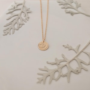 May include: A gold-colored necklace with a pendant featuring a sun and crescent moon design. The necklace is displayed on a white surface with decorative gray leaves. The pendant is round and has etched details.