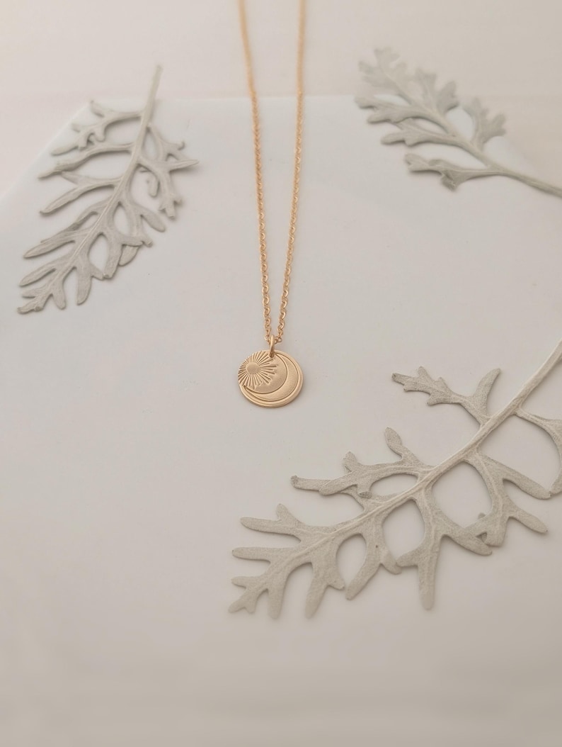 May include: A gold-colored necklace with a pendant featuring a sun and crescent moon design. The necklace is displayed on a white surface with decorative gray leaves. The pendant is round and has etched details.