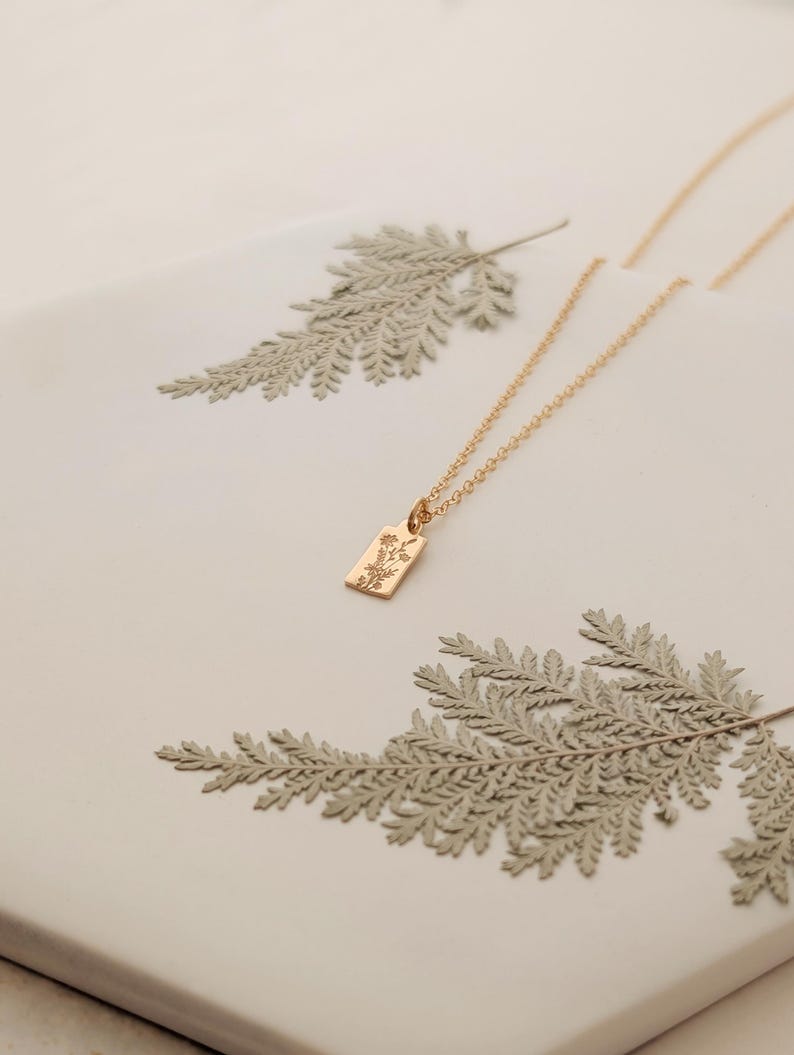 May include: A delicate gold-colored necklace with a rectangular pendant featuring a floral design. The necklace rests on a white surface, adorned with dried fern leaves. The pendant is approximately 0.5 inches in length.