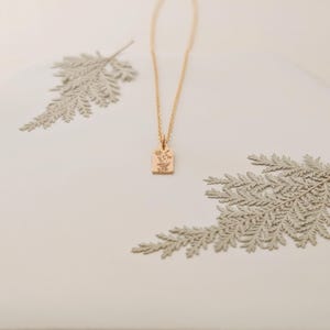 May include: A gold-colored necklace with a rectangular pendant featuring a floral design. The necklace is displayed on a white surface with decorative fern-like elements. The pendant is approximately 0.5 inches in length.